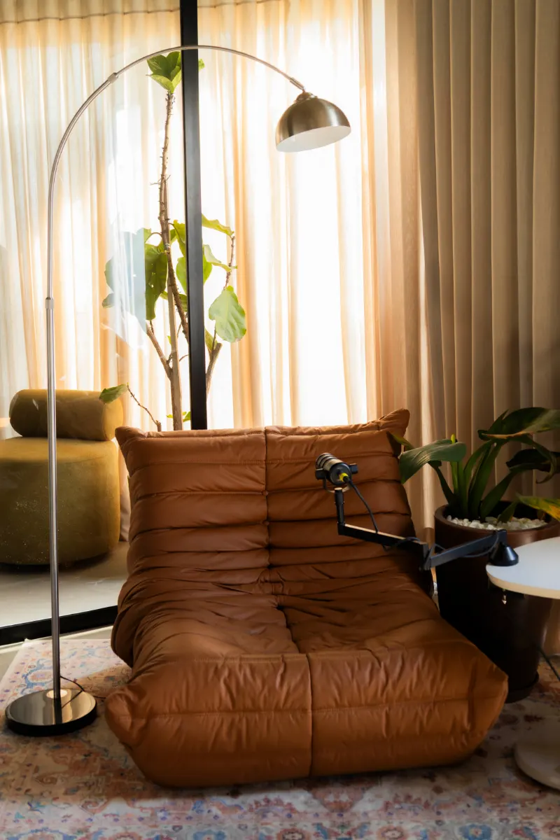Cenário Puff: poltrona puff de couro caramelo com microfone, luminária de arco, plantas e cortina clara