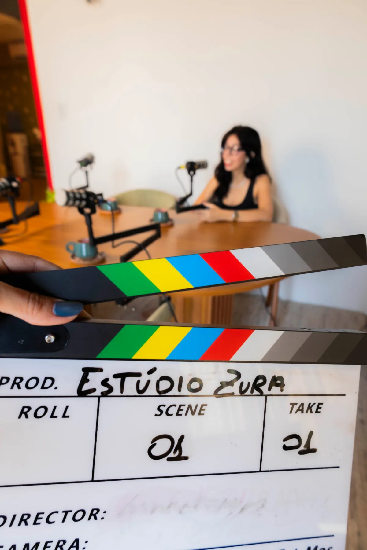 Bastidores: claquete do Estúdio Zura em primeiro plano, com a mesa de gravação de podcast ao fundo